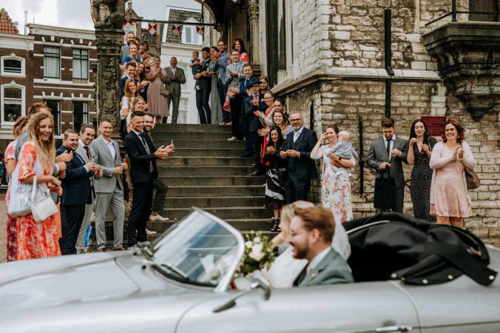 Bruidspaar komt met open auto aan bij het stadhuis van Gouda, de gasten zien hen voor het eerst en klappen enthousiast.