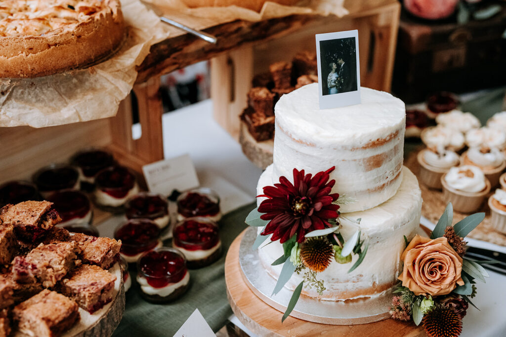 Bruidstaart met bloemen en eigengemaakt gebak daarnaast. De topper is een polaroidfoto van het bruidspaar