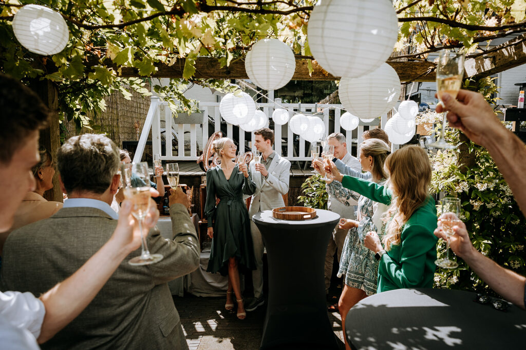 Gasten en bruidspaar proosten met champagne in eigen tuin, lampionnen hangen boven hen