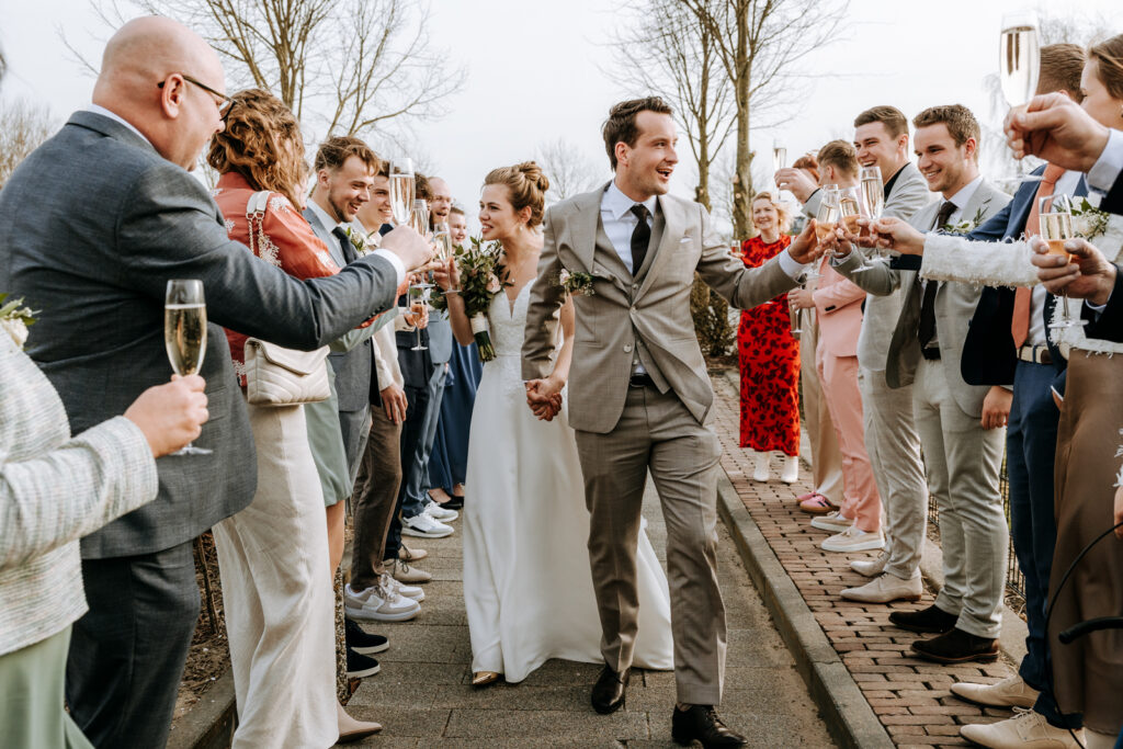 Bruidspaar in witte jurk en pak komt aan met champagne door een haag van vrienden heen die ook champagne hebben
