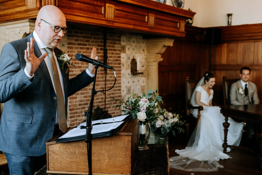 Vader van de bruid houdt toespraak tijdens trouwceremonie in oud raadhuis, bruidspaar op de achtergrond