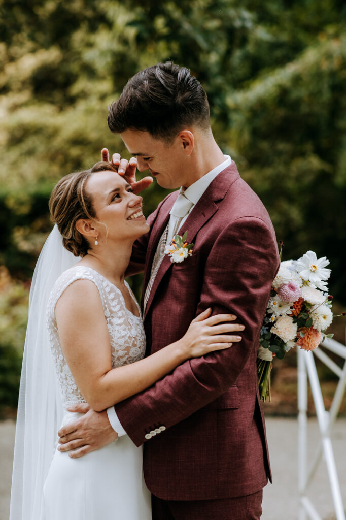 Bruid en bruidegom staan op bruggetje in park in Rotterdam, bruid kijkt op naar bruidegom en bruidegom aait gezicht bruid, gefotografeerd door trouwfotograaf in Rotterdam