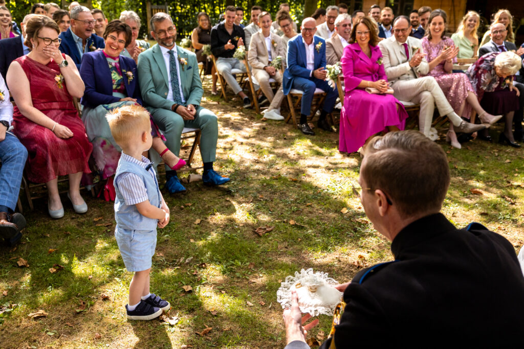 Het neefje dat de ringen brengt tijdens de buitenceremonie kijkt verlegen naar de lachende gasten