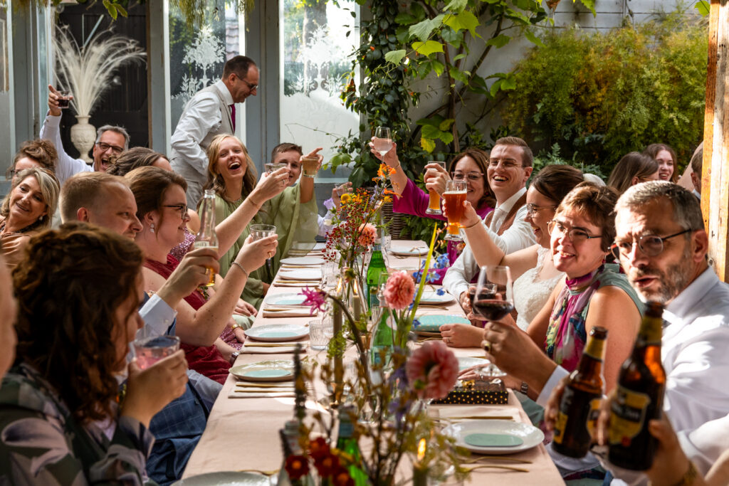 Een kleurrijke, vrolijke foto van het bruidspaar dat met alle gasten aan het diner zit en ze heffen het glas voor een proost.