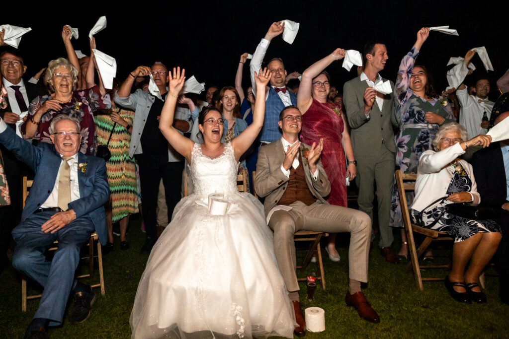 Vrolijke, kleurrijke foto van een bruidspaar met al hun gasten dat naar een grappig toneelstukje kijkt en iedereen zwaait uitgelaten met witte zaldoeken