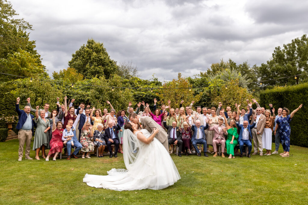 Vrolijke groepsfoto van alle bruiloftsgasten die juichen terwijl het bruidspaar elkaar kust