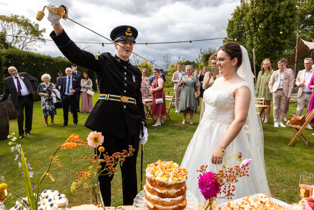 Vrolijke en kleurrrijke foto van bruid en bruidegom in uniform, die de bruidstaart gaan aansnijden met de sabel van de bruidegom.